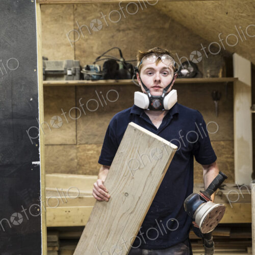 Young man wearing dust mask and protective goggles standing in a workshop, holding… Young man wearing dust mask and protective goggles standing in a workshop, holding…
