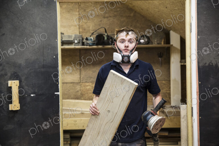 Young man wearing dust mask and protective goggles standing in a workshop, holding…