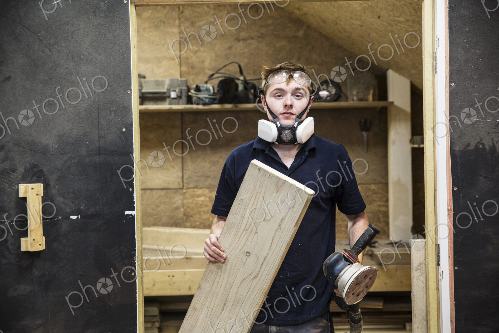 Young man wearing dust mask and protective goggles standing in a workshop, holding…