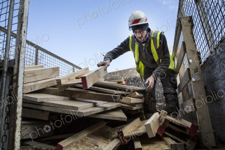 Young man wearing hard hat and reflective vest standing on top of stack of recycled wood.