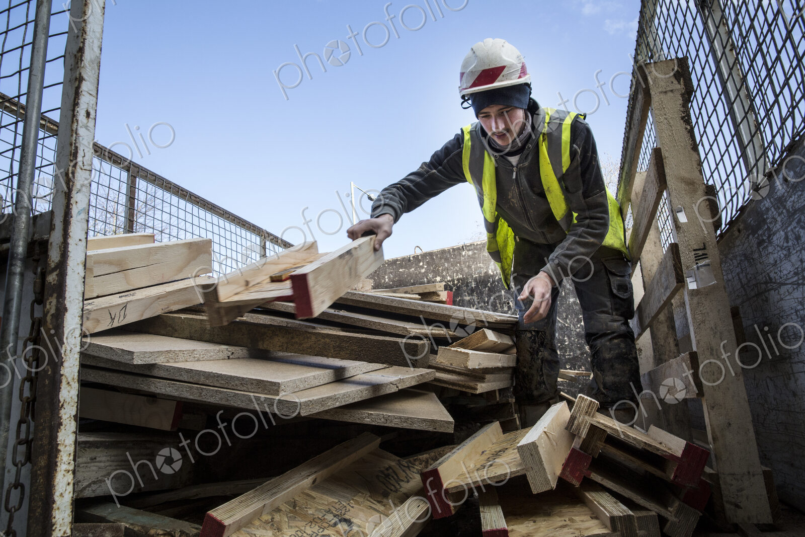 Young man wearing hard hat and reflective vest standing on top of stack of recycled wood.
