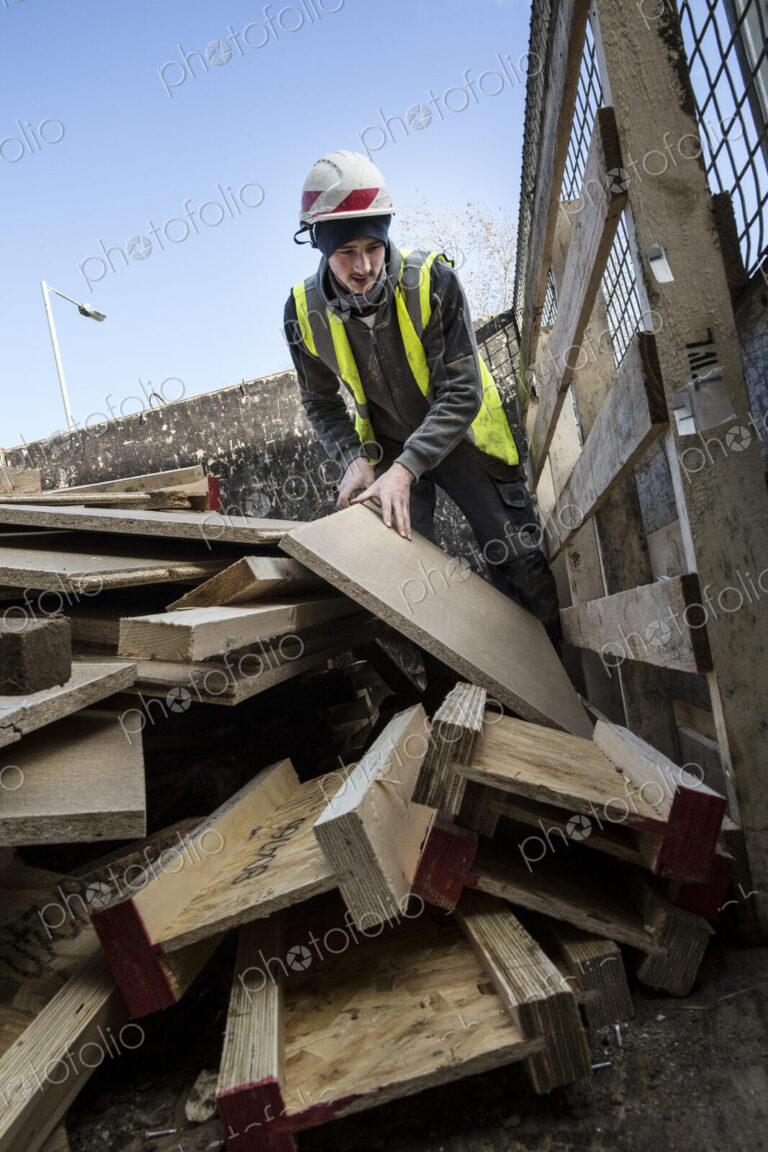 Young man wearing hard hat and reflective vest standing on top of stack of recycled wood.