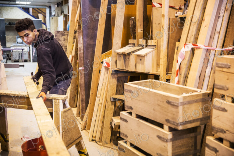 Young man wearing work gloves standing in warehouse, balancing plank of wooden on…