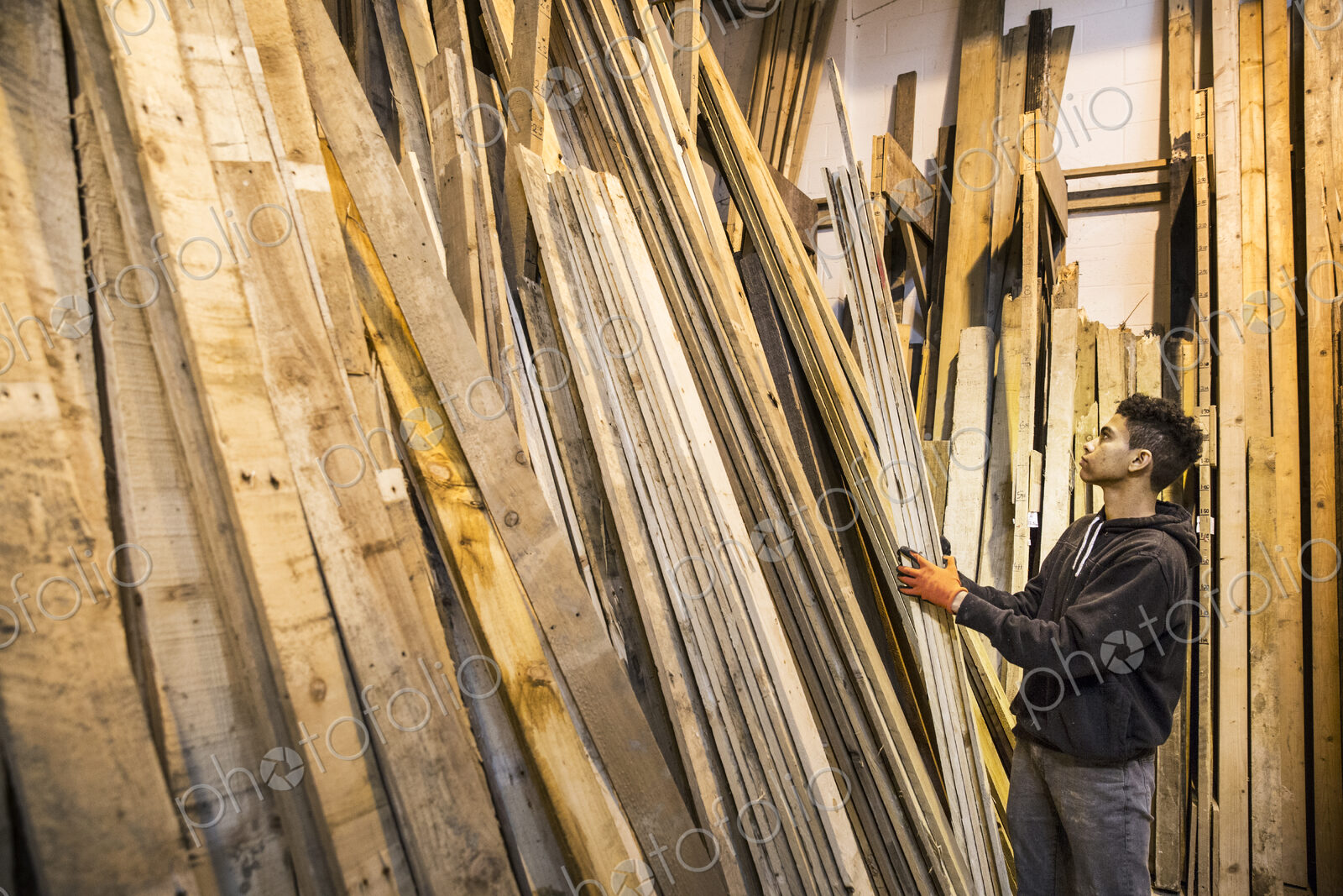 Young man wearing work gloves standing next to a stack of wooden planks in a warehouse.