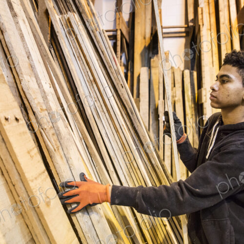Young man wearing work gloves standing next to a stack of wooden planks in a warehouse. Young man wearing work gloves standing next to a stack of wooden planks in a warehouse.