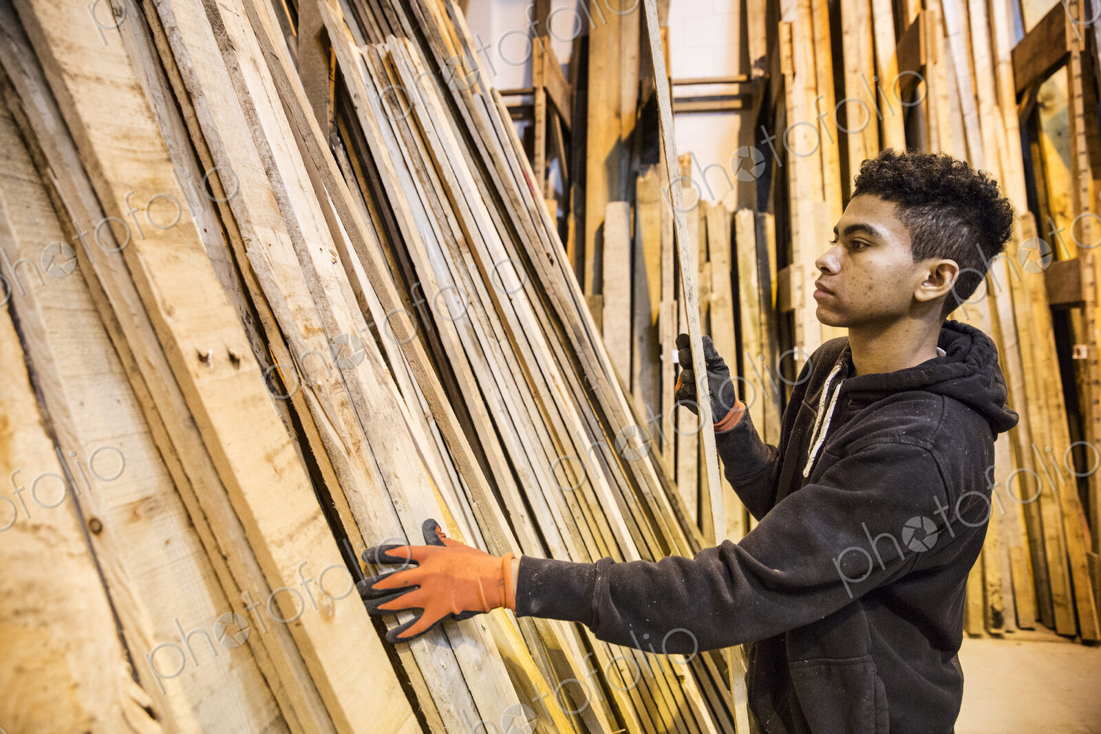 Young man wearing work gloves standing next to a stack of wooden planks in a warehouse.