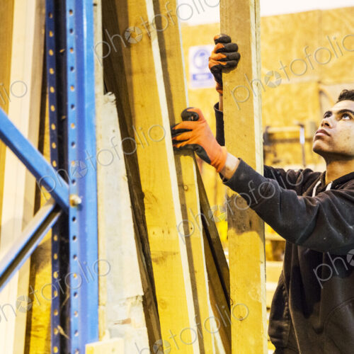 Young man wearing work gloves standing next to a stack of wooden planks in a… Young man wearing work gloves standing next to a stack of wooden planks in a…