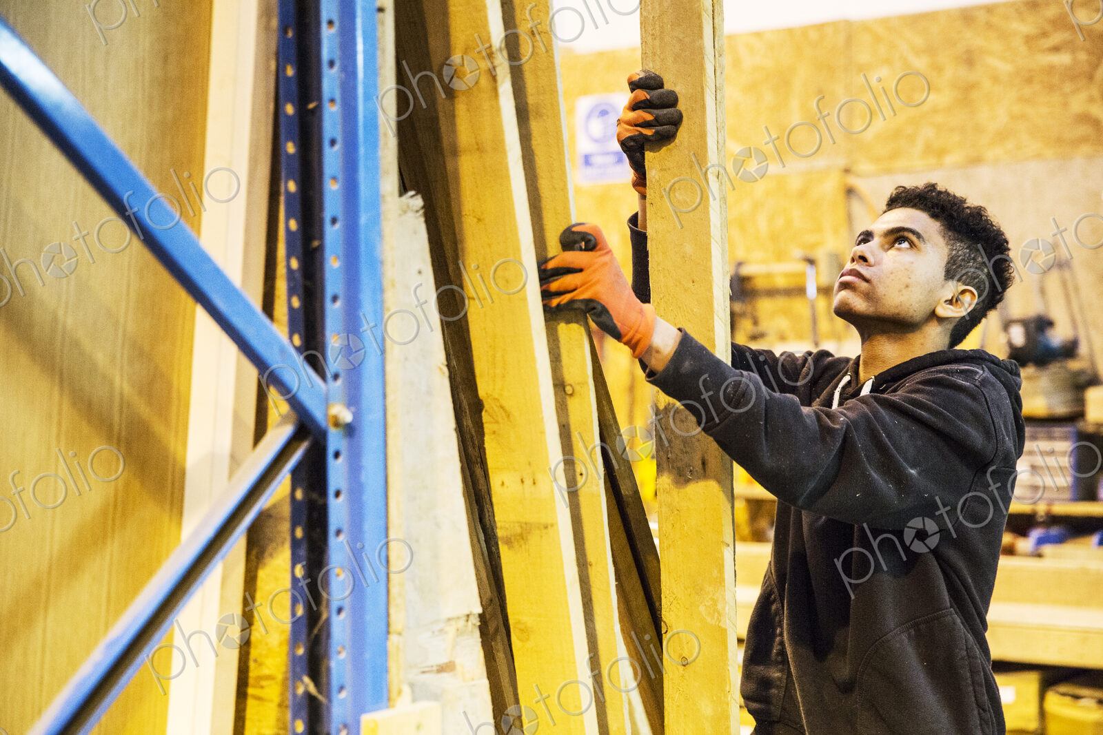 Young man wearing work gloves standing next to a stack of wooden planks in a…