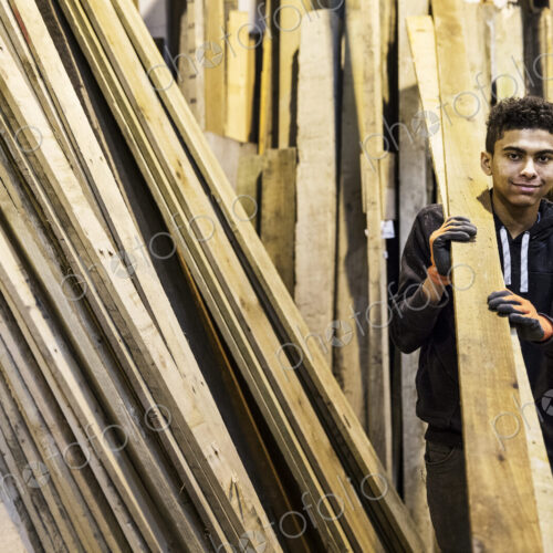 Young man wearing work gloves standing next to a stack of wooden planks in a… Young man wearing work gloves standing next to a stack of wooden planks in a…