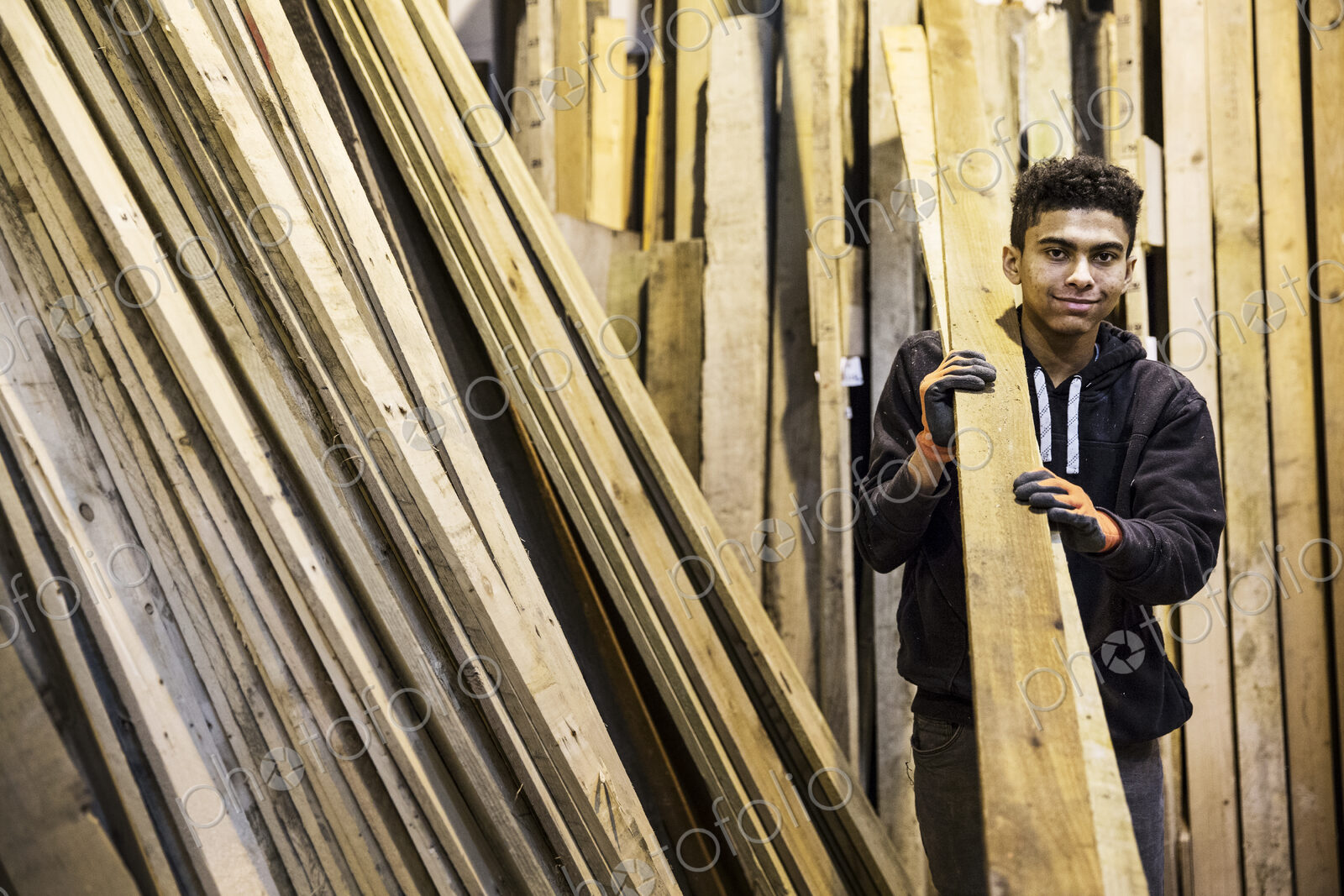 Young man wearing work gloves standing next to a stack of wooden planks in a…