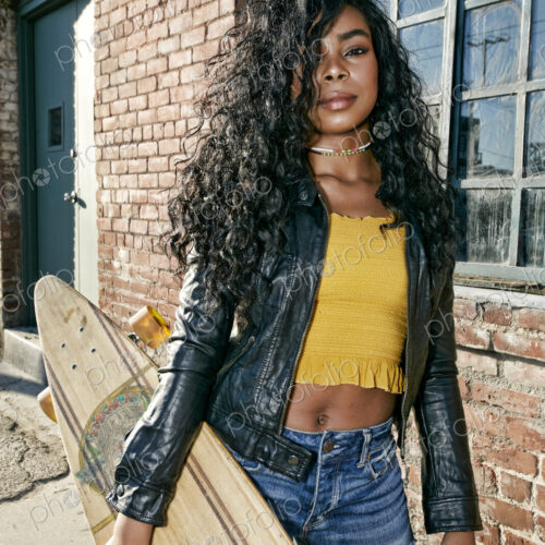 Young smiling woman with long curly black hair standing on pavement, holding…