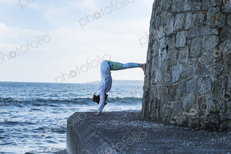 Young woman doing handstand by the ocean, with her legs against a wall.