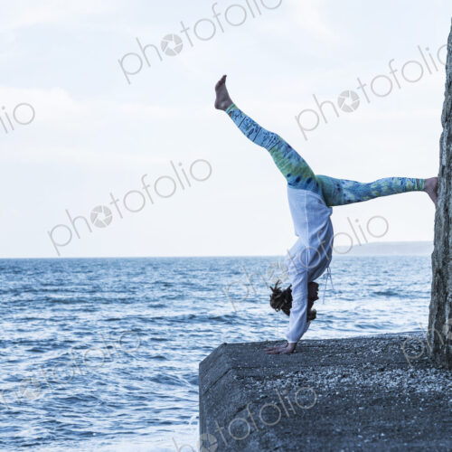 Young woman doing handstand on a wall by the ocean, one leg raised. Young woman doing handstand on a wall by the ocean, one leg raised.
