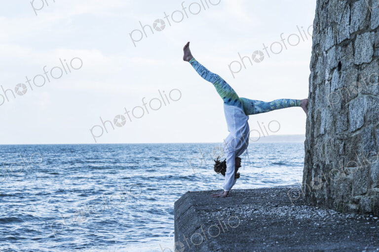 Young woman doing handstand on a wall by the ocean, one leg raised.