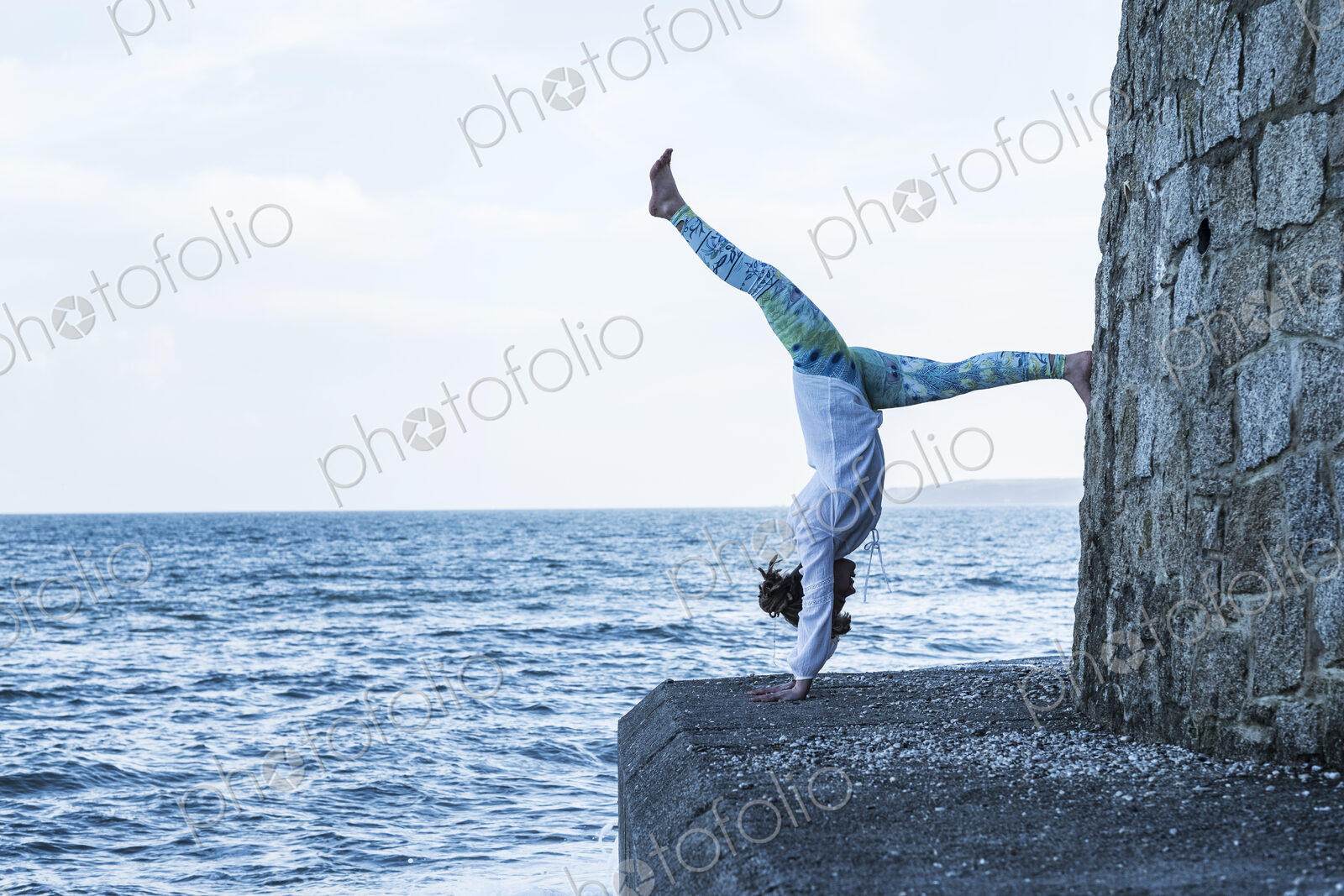 Young woman doing handstand on a wall by the ocean, one leg raised.