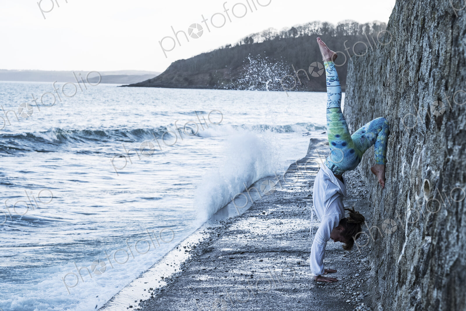 Young woman doing handstand on a wall by the ocean, one leg raised.