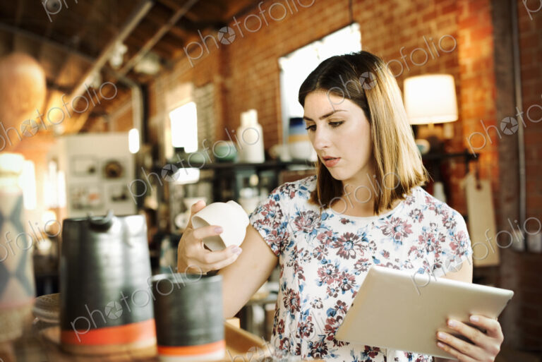 Young woman in a shop, holding a digital tablet and a ceramic mug.