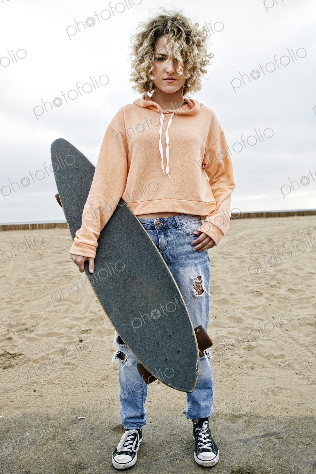 Young woman on sandy beach, holding skateboard, looking at camera.