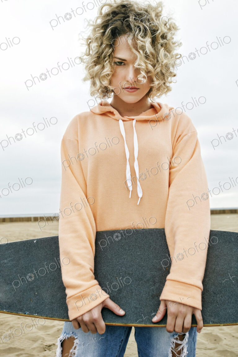 Young woman on sandy beach, holding skateboard, looking at camera.