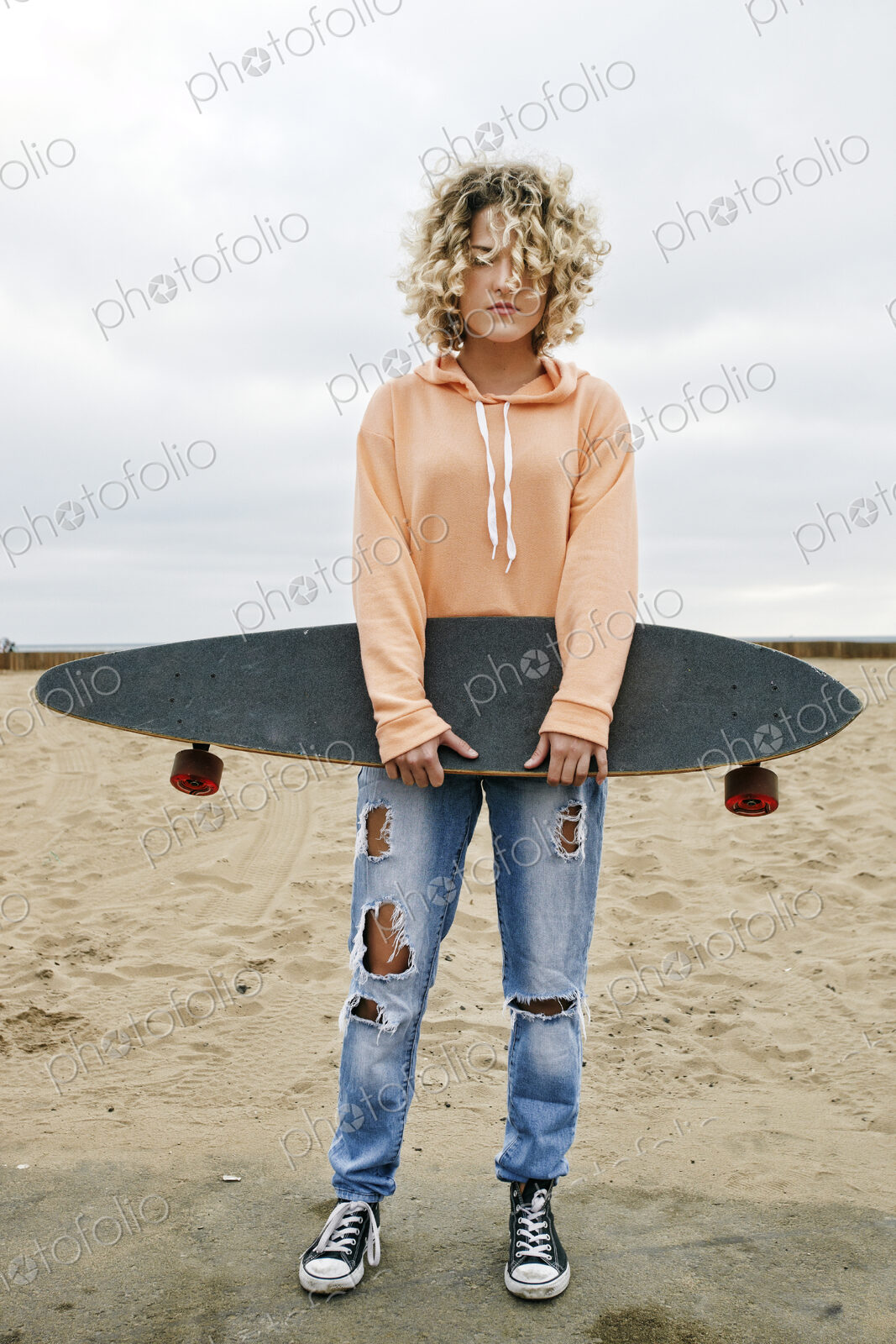 Young woman on sandy beach, holding skateboard, looking at camera.