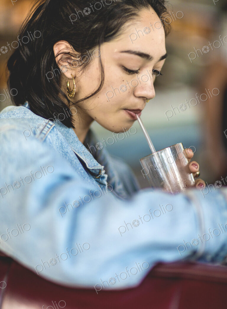 Young woman sitting at a bar counter, holding a glass, drinking with a straw.