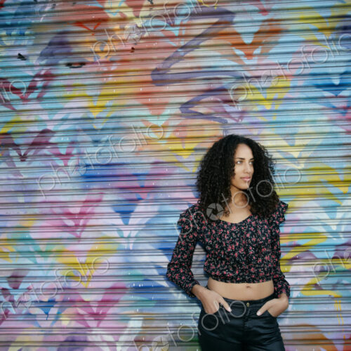 Young woman with black curly hair standing in front of shutter covered in colourful… Young woman with black curly hair standing in front of shutter covered in colourful…