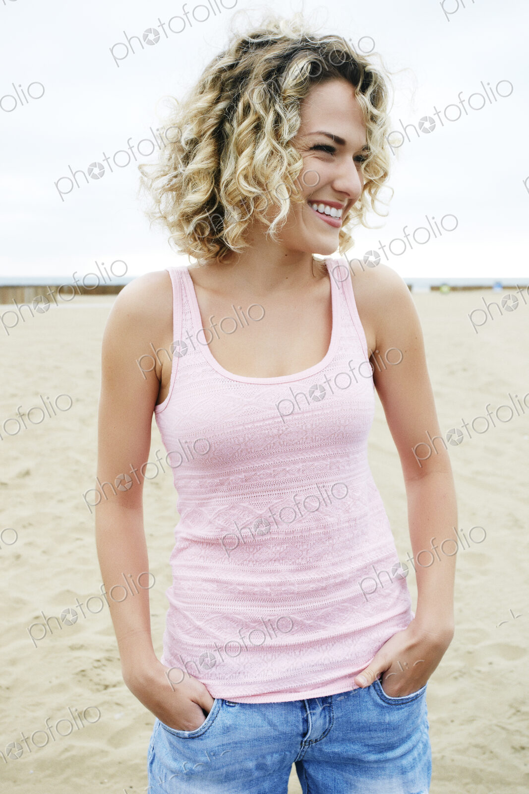 Young woman with blond curly hair standing on sandy beach.