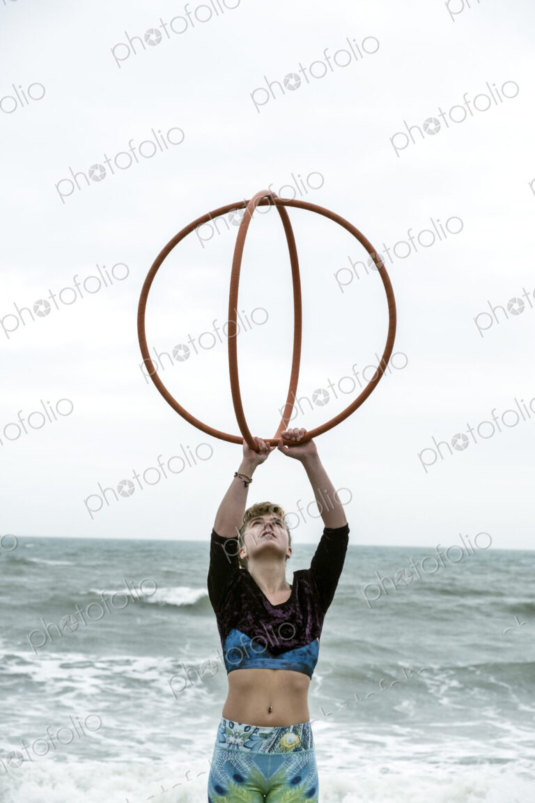 Young woman with brown hair and dreadlocks standing by the ocean, balancing two hula…