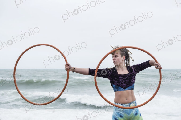 Young woman with brown hair and dreadlocks standing by the ocean, balancing two hula…