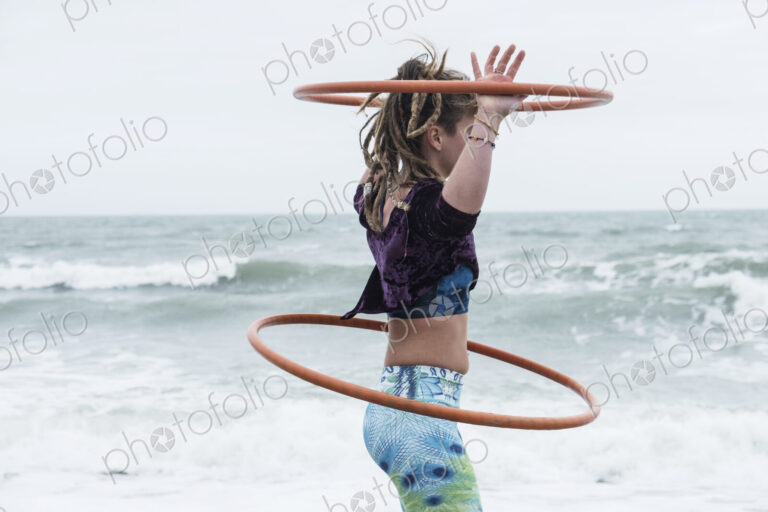 Young woman with brown hair and dreadlocks standing by the ocean, balancing two hula…
