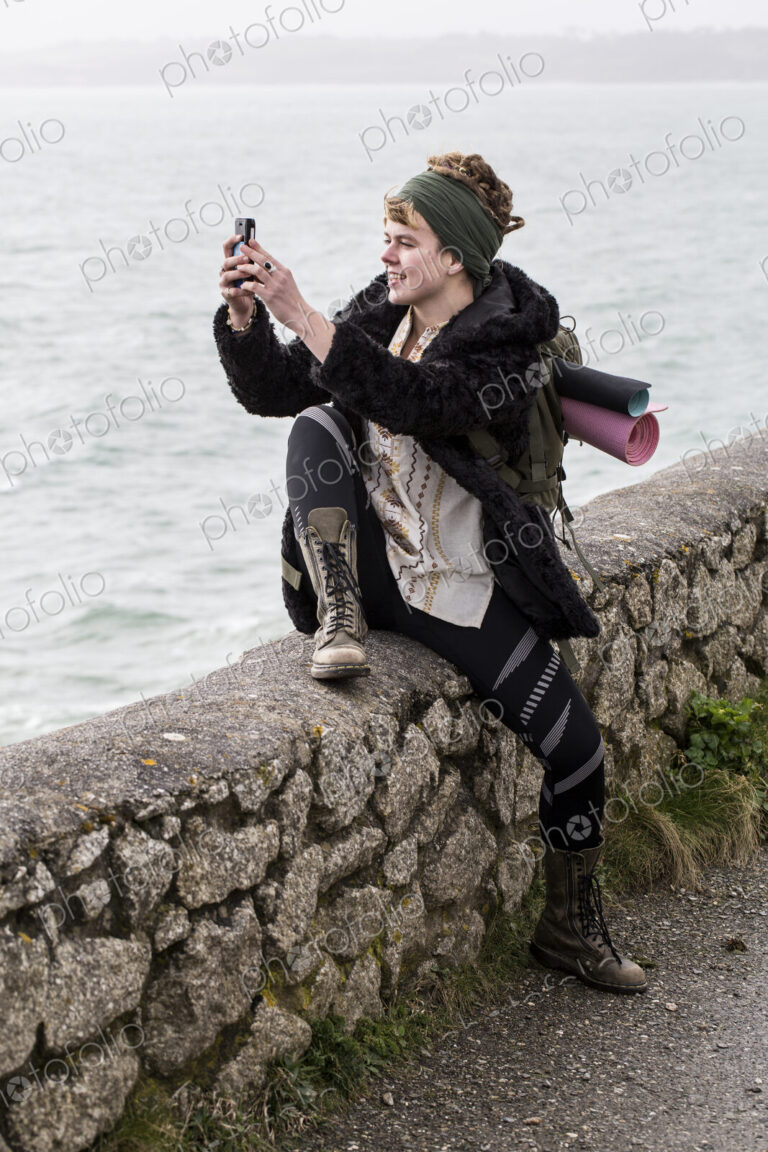 Young woman with brown hair and dreadlocks wearing headscarf sitting on dry-stone wall…