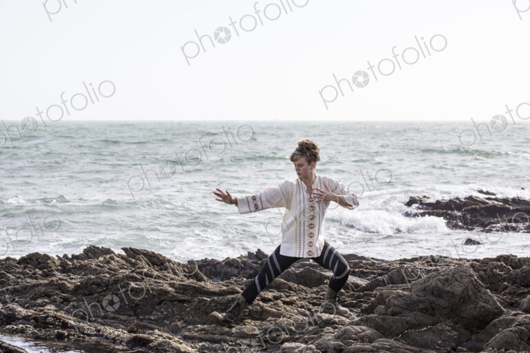 Young woman with brown hair and dreadlocks wearing white blouse standing on rocky…