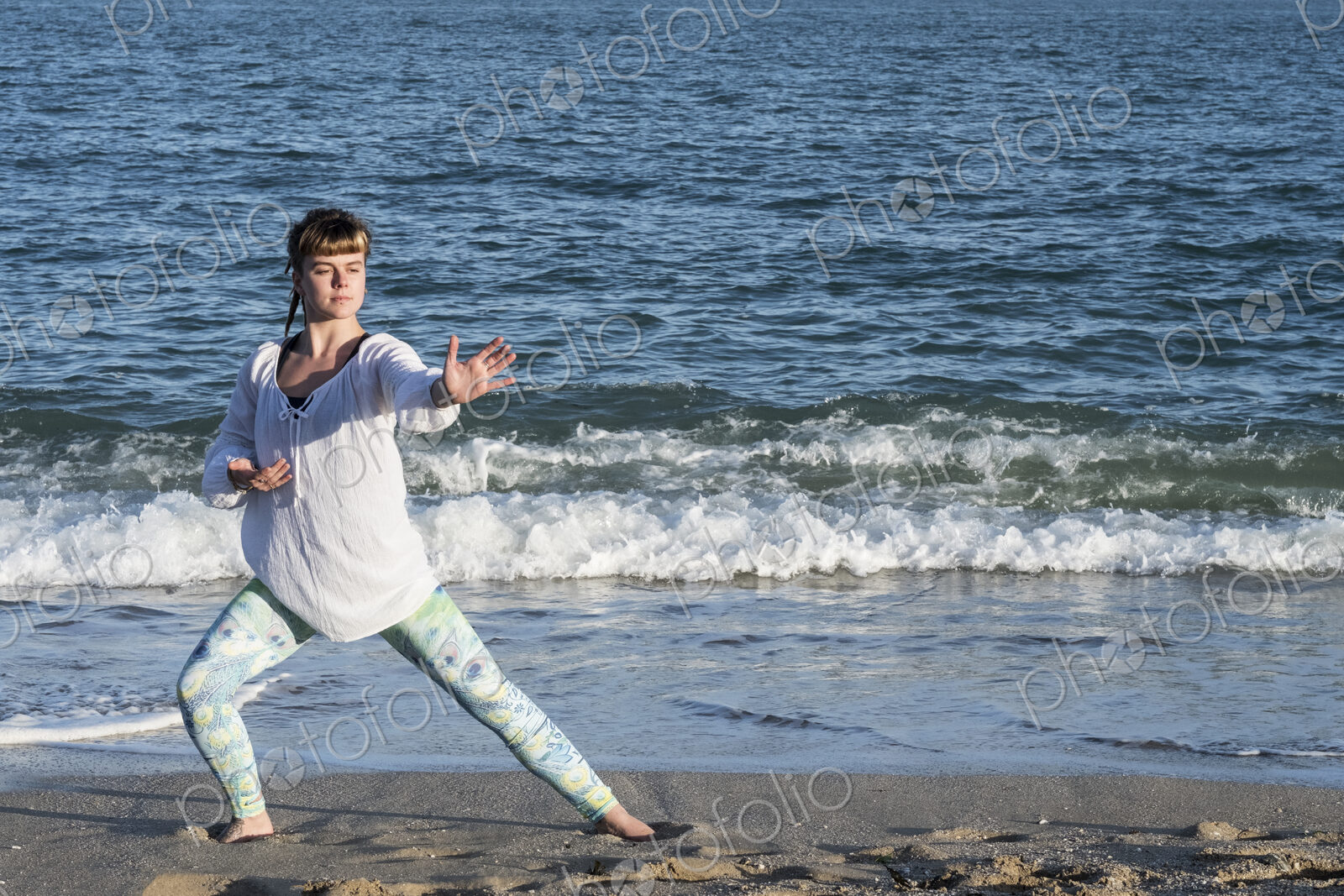 Young woman with brown hair wearing white blouse standing on a beach by the ocean,…