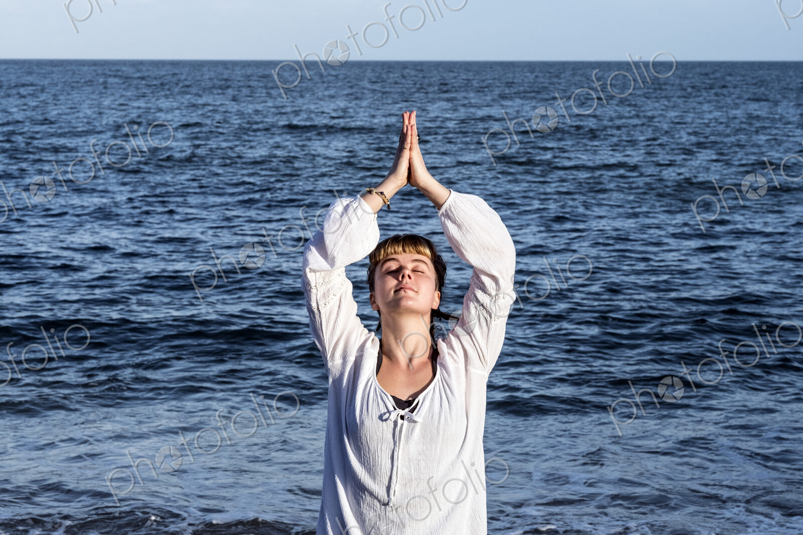 Young woman with brown hair wearing white blouse standing on a beach by the ocean,…