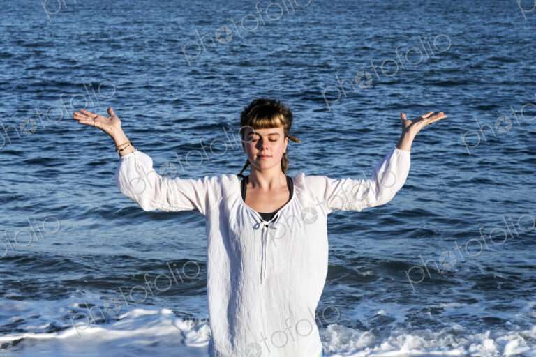 Young woman with brown hair wearing white blouse standing on a beach by the ocean,…
