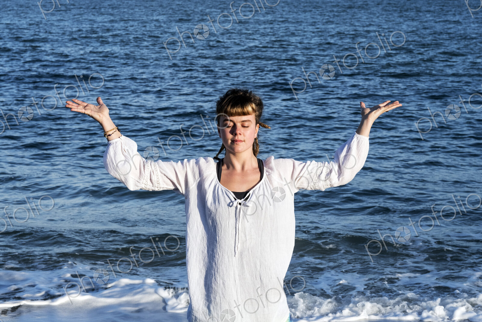 Young woman with brown hair wearing white blouse standing on a beach by the ocean,…