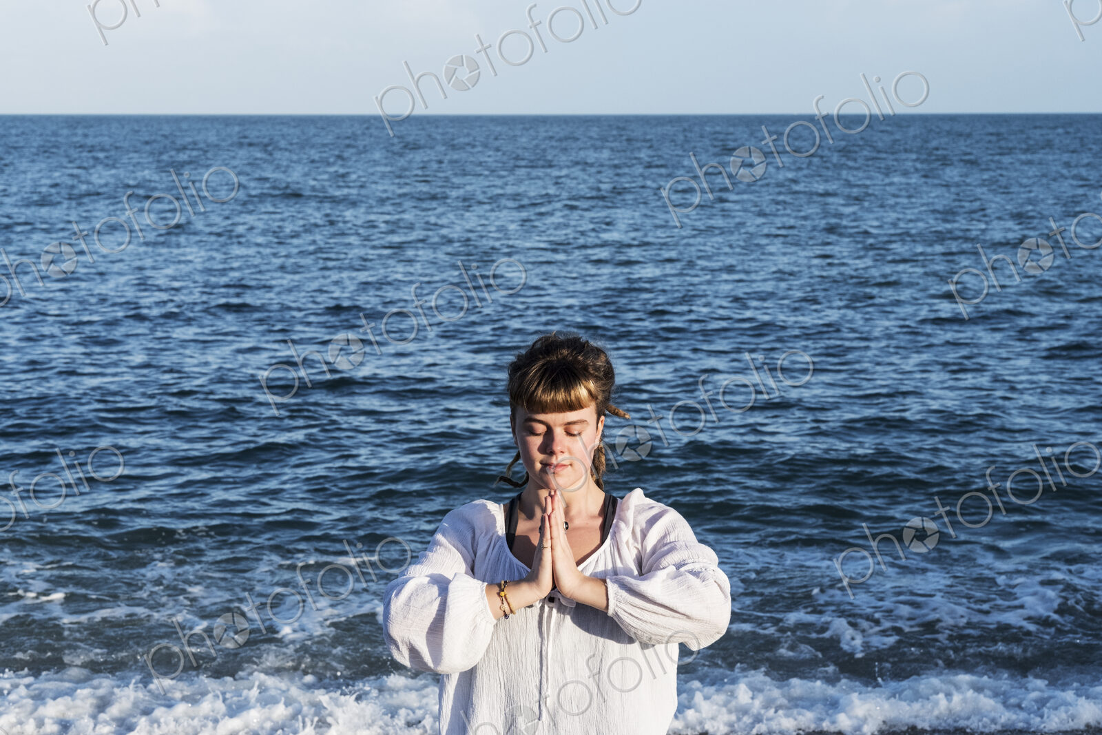 Young woman with brown hair wearing white blouse standing on a beach by the ocean,…