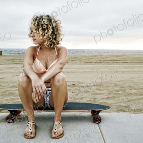 Young woman with curly blond hair wearing pink bikini sitting on skateboard on sandy… Young woman with curly blond hair wearing pink bikini sitting on skateboard on sandy…