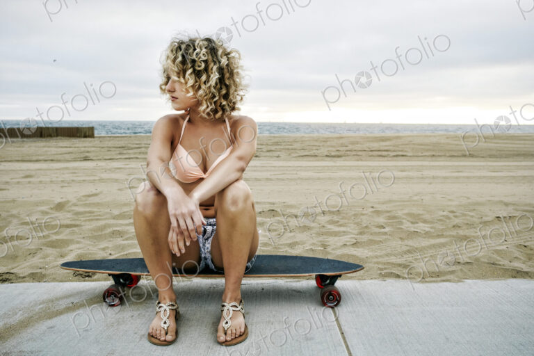 Young woman with curly blond hair wearing pink bikini sitting on skateboard on sandy…