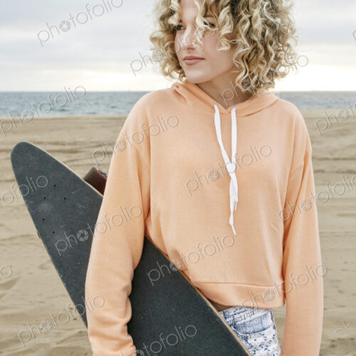 Young woman with curly blond hair wearing pink hoodie standing on sandy beach, holding… Young woman with curly blond hair wearing pink hoodie standing on sandy beach, holding…