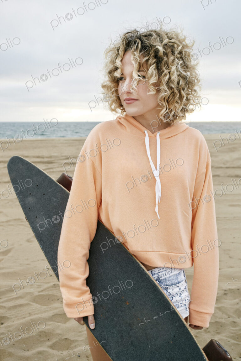 Young woman with curly blond hair wearing pink hoodie standing on sandy beach, holding…