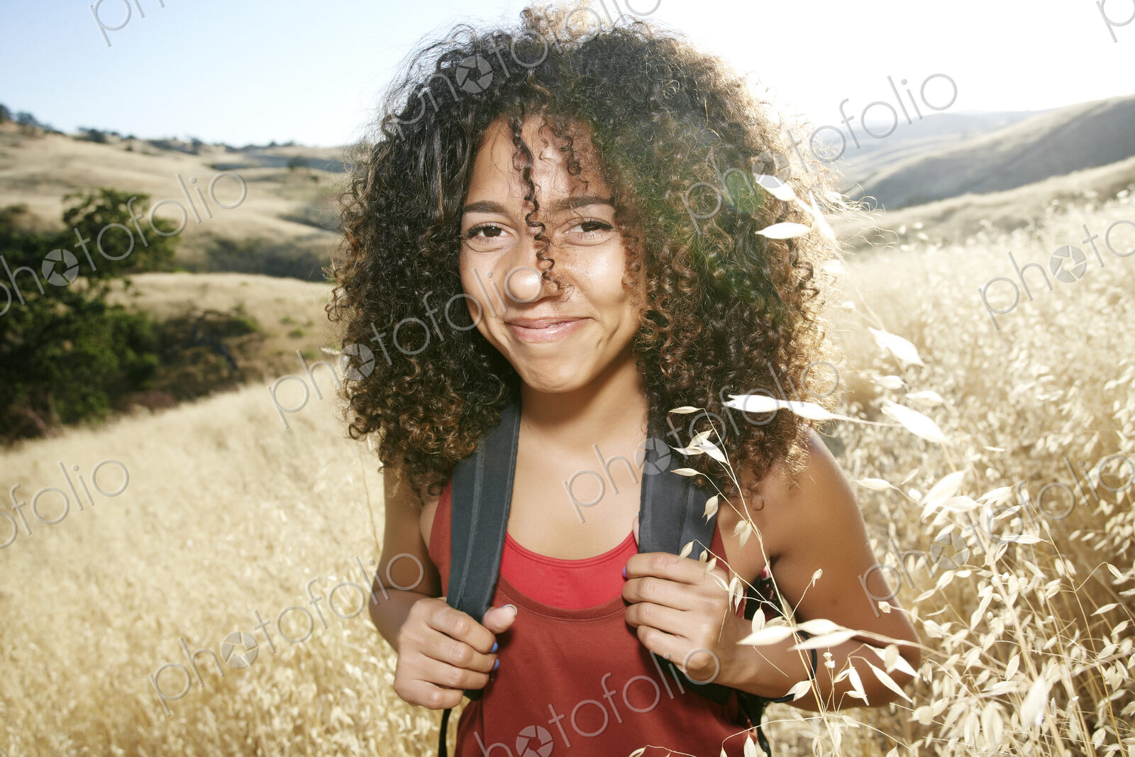 Young woman with curly brown hair hiking in urban park, smiling at camera.