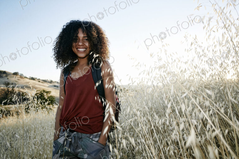 Young woman with curly brown hair hiking in urban park, smiling at camera.