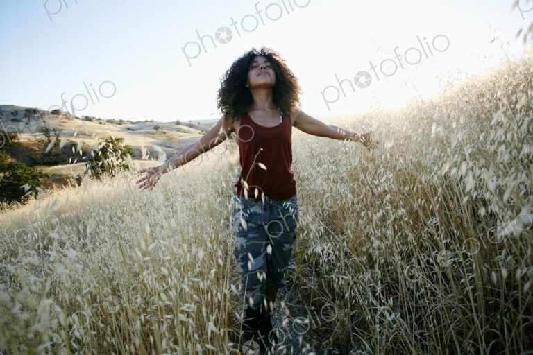 Young woman with curly brown hair hiking in urban park, standing in field, eyes closed.
