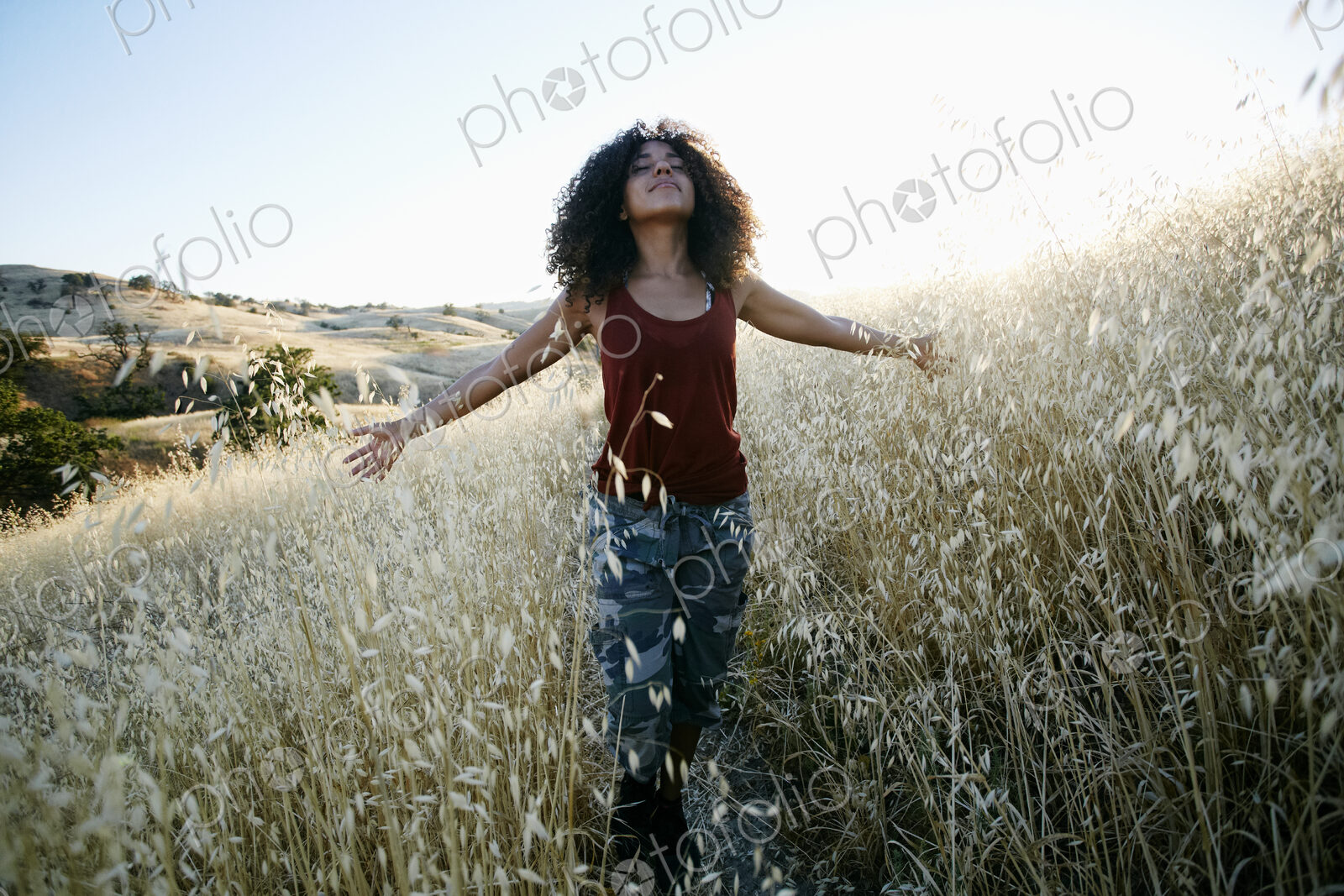 Young woman with curly brown hair hiking in urban park, standing in field, eyes closed.