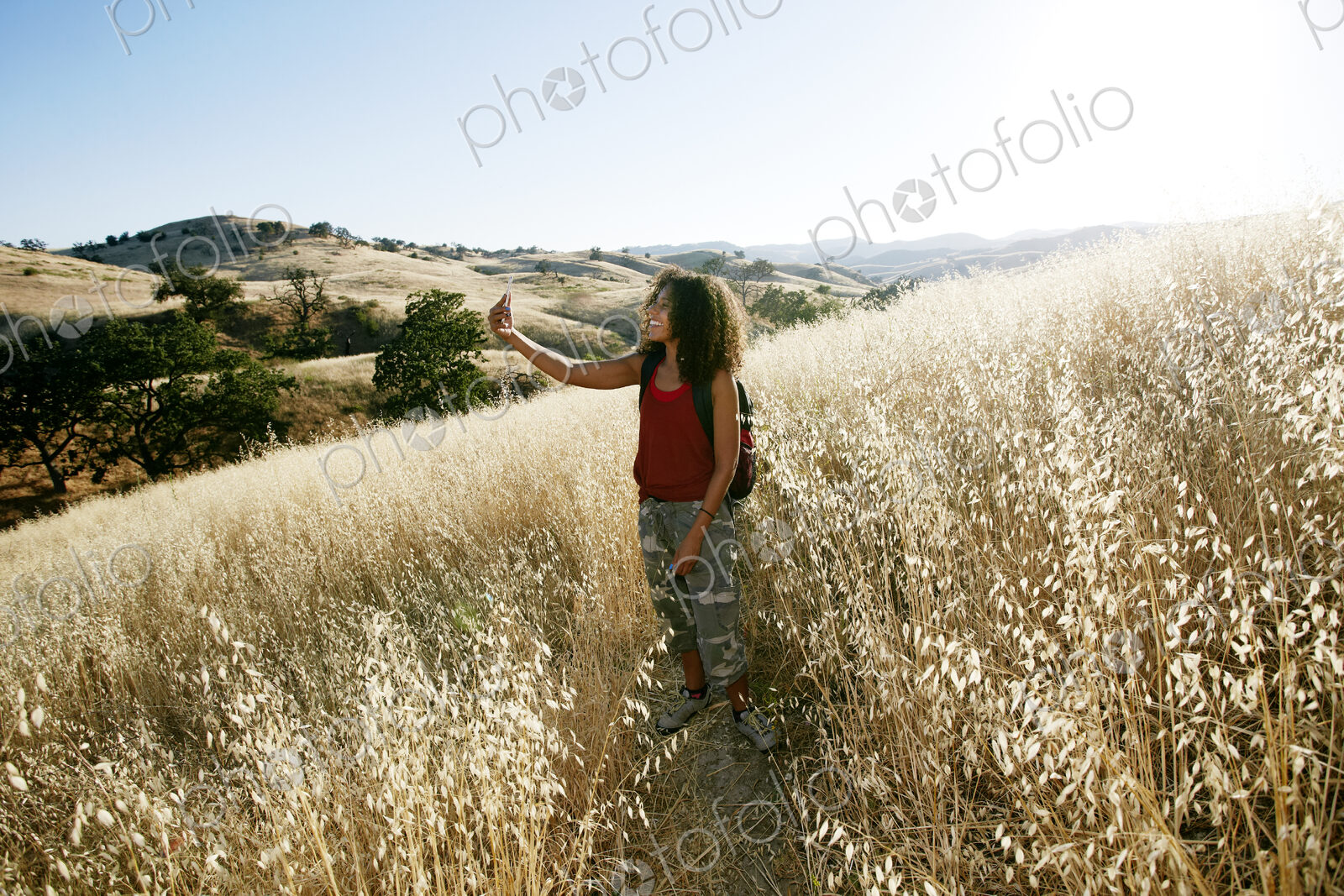 Young woman with curly brown hair hiking in urban park, taking picture with mobile phone.