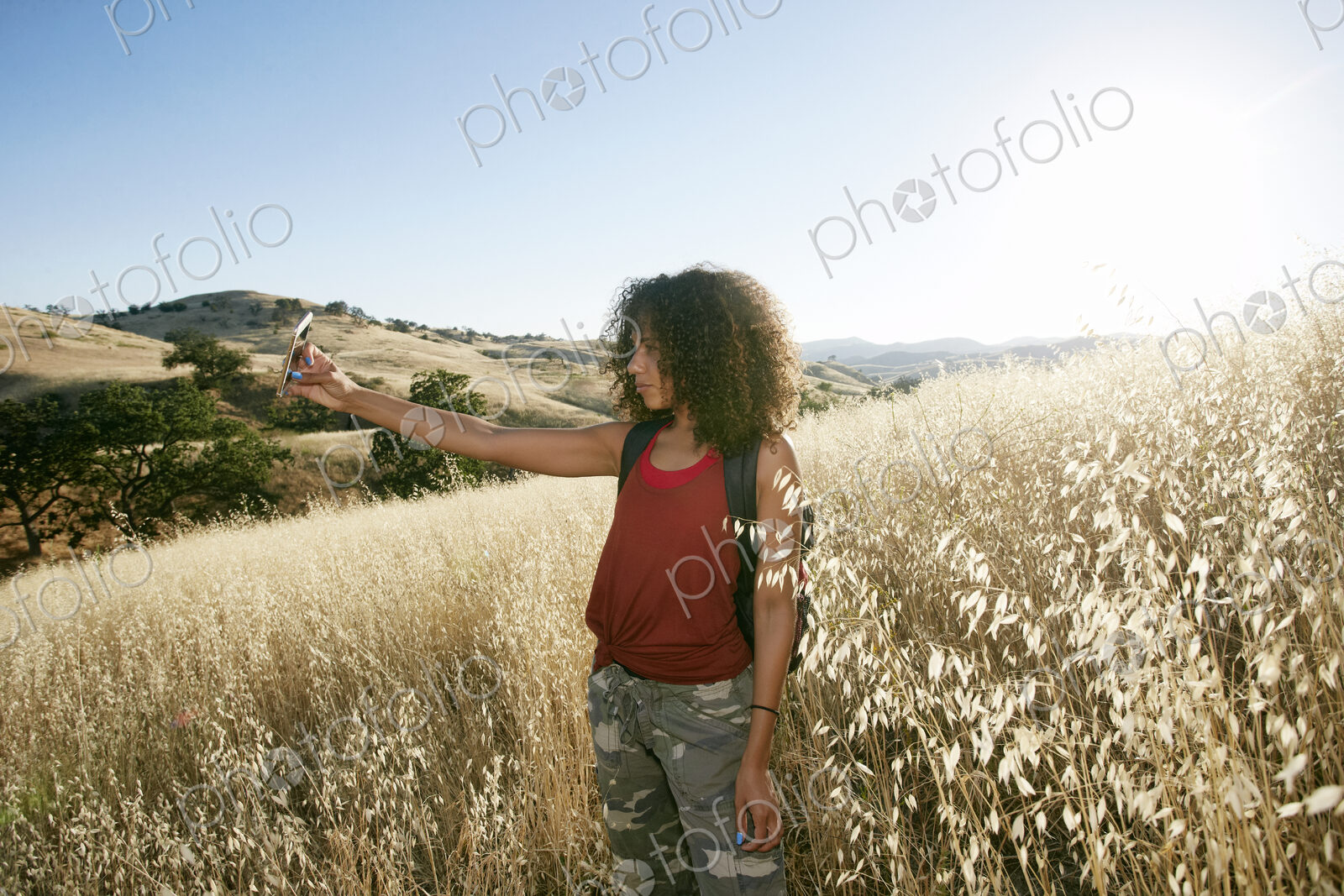 Young woman with curly brown hair hiking in urban park, taking picture with mobile phone.