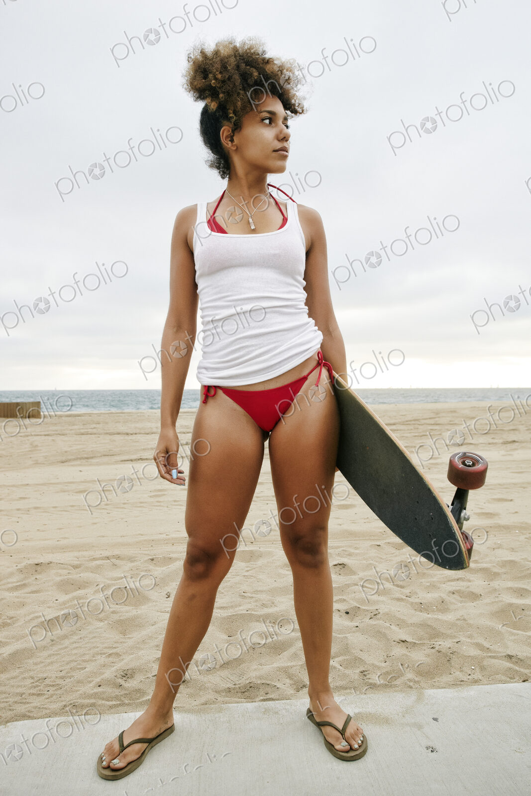 Young woman with curly brown hair wearing white vest and red bikini standing on sandy…