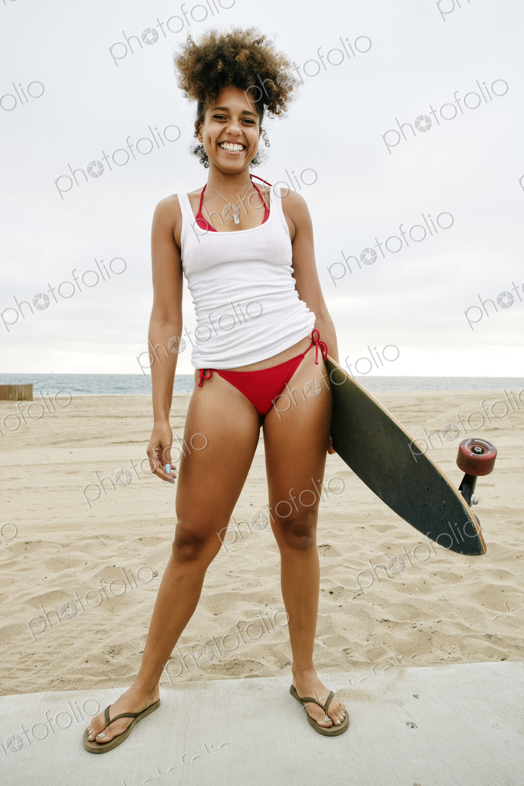 Young woman with curly brown hair wearing white vest and red bikini standing on sandy…