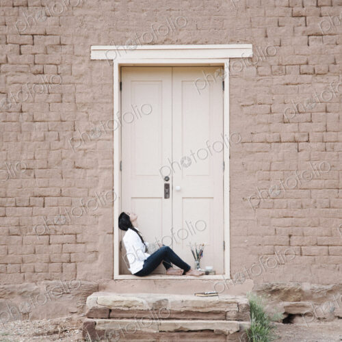 Young woman with long brown hair sitting on the ground outside the front door of a… Young woman with long brown hair sitting on the ground outside the front door of a…